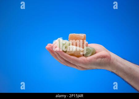 Cubetti di delizia turca colorata tenuti in mano maschile. Primo piano ripresa in studio, isolata su sfondo blu. Foto Stock