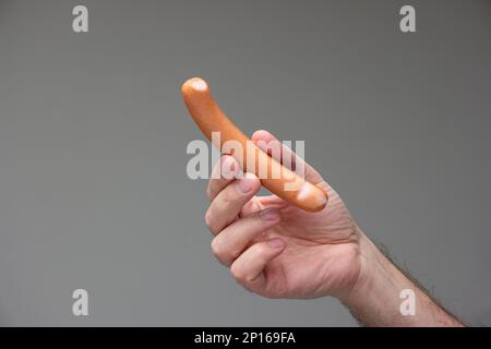 Mano maschio caucasica che tiene un cane bratwurst tra le dita isolato su sfondo grigio. Foto Stock