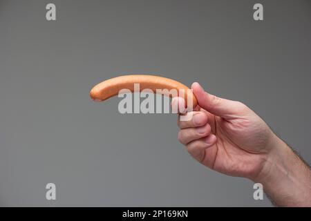 Mano maschio caucasica che tiene un cane bratwurst tra le dita isolato su sfondo grigio. Foto Stock