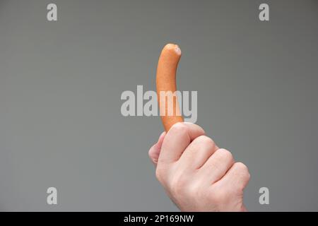 Mano maschio caucasica che tiene un cane bratwurst tra le dita isolato su sfondo grigio. Foto Stock