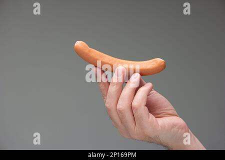 Mano maschio caucasica che tiene un cane bratwurst tra le dita isolato su sfondo grigio. Foto Stock