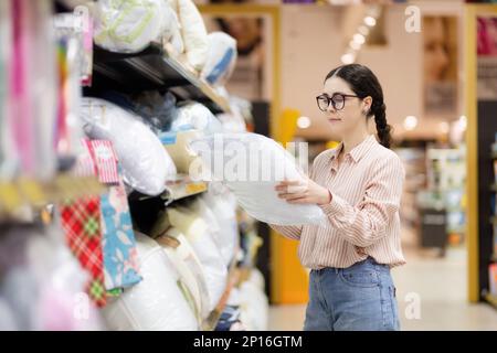 Donna caucasica con bicchieri sceglie cuscini e biancheria da letto. Tessili per la casa in supermercato. Shopping. Foto Stock