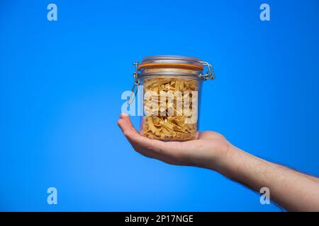 Vasetto di vetro con coperchio ripieno di pasta italiana fatta in casa tenuto in mano da maschio caucasico isolato su sfondo blu shot studio. Foto Stock