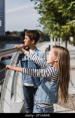 eleganti bambini in jeans giubbotti in piedi vicino a recinzione metallica sul fiume, immagine stock Foto Stock