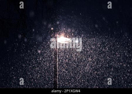 Un ritratto di una luce stradale accesa o di una lanterna, che illumina una strada durante una tempesta di neve di notte. Tutti i fiocchi di neve sono visibili nella trave di l Foto Stock