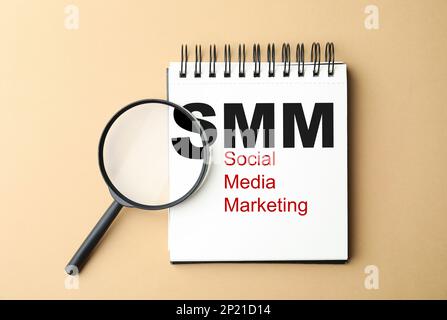 Notebook con testo SMM (Social Media Marketing) e lente di ingrandimento su sfondo beige, piatto Foto Stock