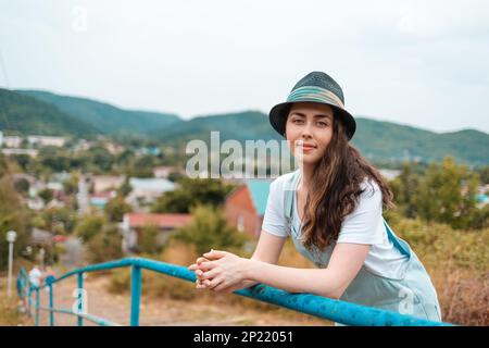 Ritratto di una giovane bella bruna donna in cappello in posa vicino alla ringhiera contro una vista rurale. Foto Stock