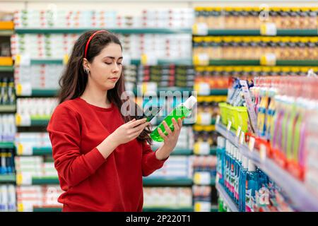 Una giovane donna caucasica scansiona il codice qr su una bottiglia di shampoo usando il suo smartphone. Sullo sfondo c'è un supermercato. Concetto di tecnologie moderne Foto Stock