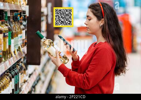 Una giovane donna caucasica esegue la scansione del codice qr su una bottiglia di vino utilizzando il suo smartphone. Icona del codice QR sopra il prodotto. Sullo sfondo, mensole con bevanda Foto Stock