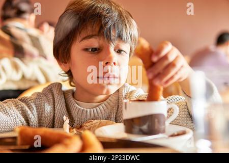 bambino di età elementare che immergerà i churros in una tazza di cioccolato caldo Foto Stock