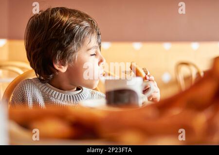 bambino di età elementare che mangia i churros yummy in una tazza di cioccolato caldo Foto Stock