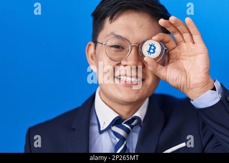 Giovane asiatico che tiene moneta virtuale bitcoin che copre l'occhio sorridente con un sorriso felice e fresco sul viso. visualizzazione dei denti. Foto Stock