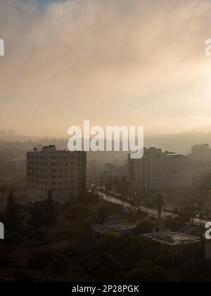 Il paesaggio urbano di Ramallah all'alba di fronte al sole al tramonto, agli edifici alti, al viale, agli alberi e ai raggi di luce volumetrici Foto Stock