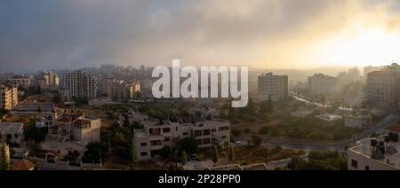 Ramallah, Ramallah e al-Bireh Governatorato, Palestina, 12 luglio 2022: Ramallah paesaggio urbano all'alba con tramonto, alti edifici e alberi di fronte al sole Foto Stock