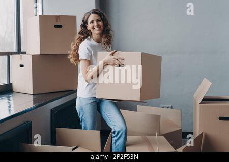 Eccitato latina femmina proprietario di abitazione inquilino che trasporta la scatola di cartone con le cose personali sul giorno commovente. Donna felice imballaggio oggetti per il trasferimento a nuovo Foto Stock