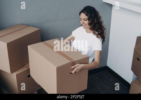 Sorridente proprietario di una casa donna ispanica affitolare in possesso di una scatola di cartone imballato con oggetti personali, in movimento in un nuovo appartamento di casa. Ragazza felice porta le cose Foto Stock