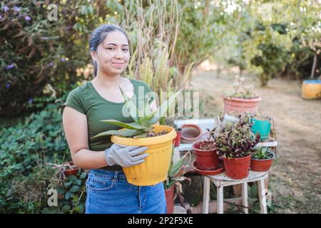 Giovane contadina latina che tiene un vaso di fiori con aloe vera pianta nel suo giardino biologico Foto Stock