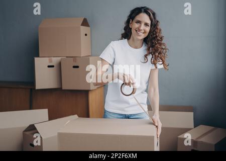 Donna spagnola sorridente imballaggio roba in scatola di cartone, utilizzando nastro adesivo guardando la macchina fotografica. Confezione di cartone sigillante femmina con nastro adesivo, pr Foto Stock