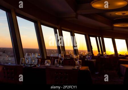 Un bagliore arancione da un tramonto che scorre attraverso grandi finestre in un ristorante deserto con tavoli allestiti con bicchieri da bere Foto Stock