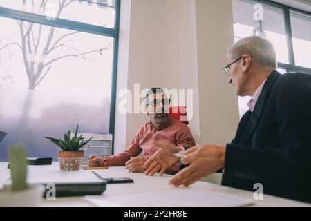Due uomini d'affari più anziani che hanno una conversazione mentre sono seduti alla scrivania presso l'area di co-lavoro Foto Stock
