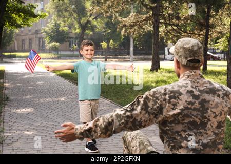 Ragazzino con bandiera degli Stati Uniti che corre verso il padre in uniforme militare all'aperto. Ricongiungimento familiare Foto Stock