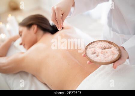 Massaggiatrice che applica il corpo di sale minerale scrub sulla parte posteriore della giovane donna Foto Stock