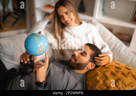 Sorridente donna europea millenaria e maschio arabo riposano nel tempo libero, guardando il globo, scegliendo il paese Foto Stock