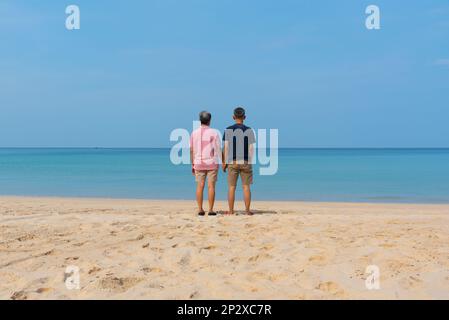 Un paio di adulti asiatici anziani che tengono le mani e si levano in piedi sulla splendida spiaggia e sullo sfondo blu del cielo. Foto Stock