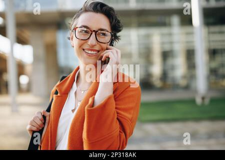 Donna d'affari sorridente che parla al telefono mentre si va in ufficio in città. Donna d'affari matura che fa i programmi per il giorno mentre cammina a. Foto Stock