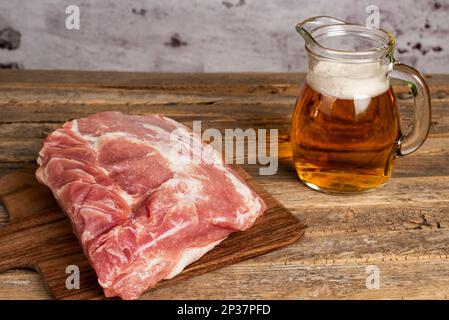 Pezzo di lombo di maiale su un tagliere accanto a una tazza di birra, elementi principali del piatto Tenderloin alla birra, su un vecchio tavolo di legno. Foto Stock