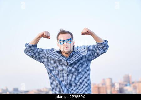 Emozione superpotenza o concetto di pugni pompati. Adorabile uomo caucasico in camicia da notte e maschera blu supereroe con gesto di forza e faccia seria. Immagini di alta qualità Foto Stock