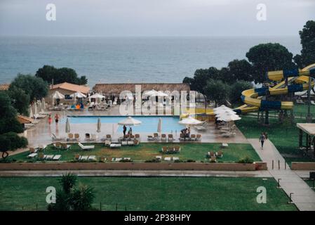 Offuscata dell'area dell'hotel con piscina e scivolo d'acqua durante la pioggia. I lettini sono vuoti, le persone fuggono dalla pioggia sotto la tettoia del Beach Cafe. Concetto Foto Stock