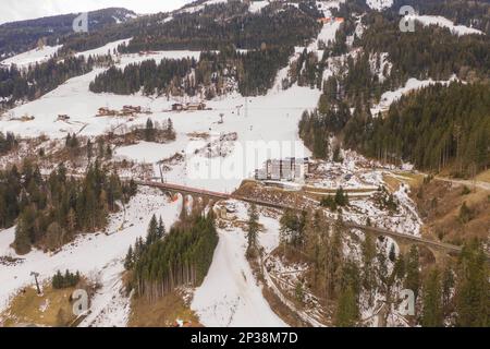 Fotografia in drone di pista del cielo di montagna, ponte ferroviario che va sopra la pendenza, treno che guida sopra il ponte e gli impianti di risalita del cielo durante la nuvolosa giornata invernale Foto Stock