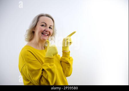 Donna in lattice giallo guanti e abiti luminosi punta a uno spazio vuoto per la pubblicità il testo di una società di pulizia sincero sorriso ristretto verde occhi come chiamare White studio Foto Stock