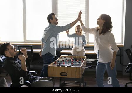 Felici amici d'affari entusiasti che giocano a calcio balilla, dando alti cinque Foto Stock