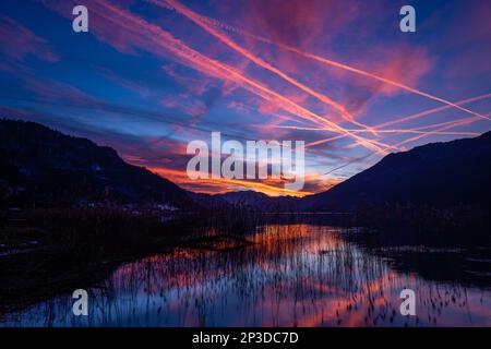 Le colorate piste di vapore degli aerei piangono-attraversano il cielo ardente mentre il sole tramonta catturato dalle riflessioni sul lago Ossiach nel Salzkammergut austriaco Foto Stock