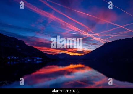 Le colorate piste di vapore degli aerei piangono-attraversano il cielo ardente mentre il sole tramonta catturato dalle riflessioni sul lago Ossiach nel Salzkammergut austriaco Foto Stock