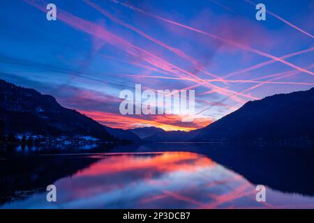 Le colorate piste di vapore degli aerei piangono-attraversano il cielo ardente mentre il sole tramonta catturato dalle riflessioni sul lago Ossiach nel Salzkammergut austriaco Foto Stock