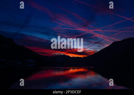 Le colorate piste di vapore degli aerei piangono-attraversano il cielo ardente mentre il sole tramonta catturato dalle riflessioni sul lago Ossiach nel Salzkammergut austriaco Foto Stock