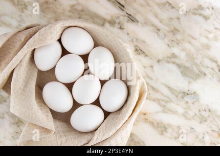 Le uova bianche di pollo si trovano in un cestino coperto con un asciugamano beige su un tavolo di marmo. Vista dall'alto della composizione pasquale, spazio per il testo Foto Stock