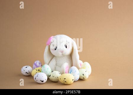 Un coniglio di Pasqua fatto di cioccolato bianco siede tra uova colorate su uno sfondo beige. Molto spazio vuoto Foto Stock