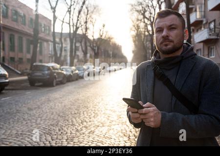 Uomo con telefono cellulare in piedi su una strada vuota in città e guardando lontano. Foto Stock
