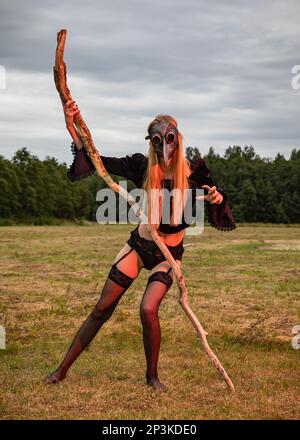 Una giovane donna vestita di nero e con una maschera nera corvata tiene un grande ramo di albero secco e si posa in mezzo a un campo in campagna Foto Stock