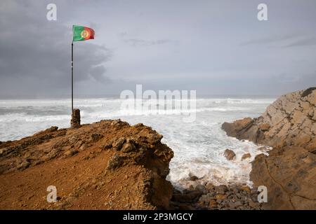 Bandiera portoghese che vola al vento con onde tempestose dell'Oceano Atlantico che si infrangono sulla spiaggia di Praia do Guincho Foto Stock
