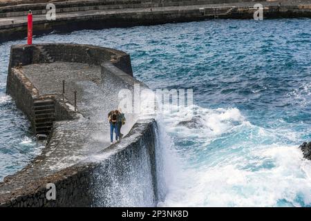 Due persone si bagnano a causa di un'onda che si schianteggia all'ingresso del porto e al molo di pesca a Puerto de la Cruz, nel nord di Tenerife, Isole Canarie. Foto Stock