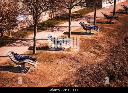 Villach, Austria - 23 febbraio 2023: Passeggiata per la città, persone che riposano su sedie a sdraio di legno lungo le rive del fiume Drau Foto Stock