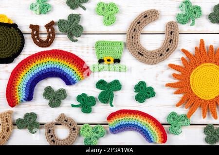 Concetto del giorno di San Patrizio. Composizione a maglia di un cappello verde, una pentola d'oro, un ferro di cavallo, un sole arancione, un arcobaleno e shamrock verde su un bosco bianco Foto Stock