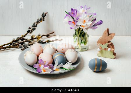 Piatto grigio con uova di Pasqua dipinte e coniglietto giocattolo in legno, fiori di cocco su tavolo bianco. Biglietto di auguri di Pasqua. Foto Stock