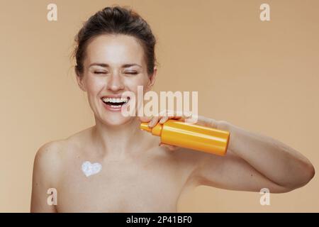 donna sorridente moderna di 40 anni con vasetto crema cosmetica e crema a forma di cuore sul petto isolato su sfondo beige. Foto Stock