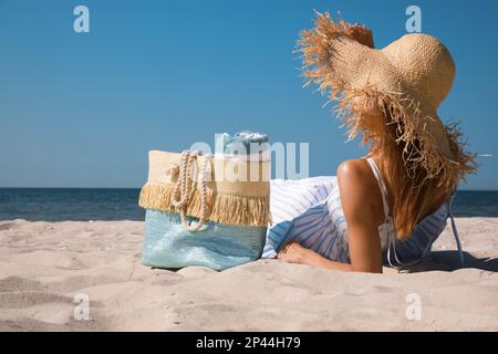 Donna con borsa da spiaggia e cappello di paglia sdraiato sulla sabbia vicino al mare Foto Stock